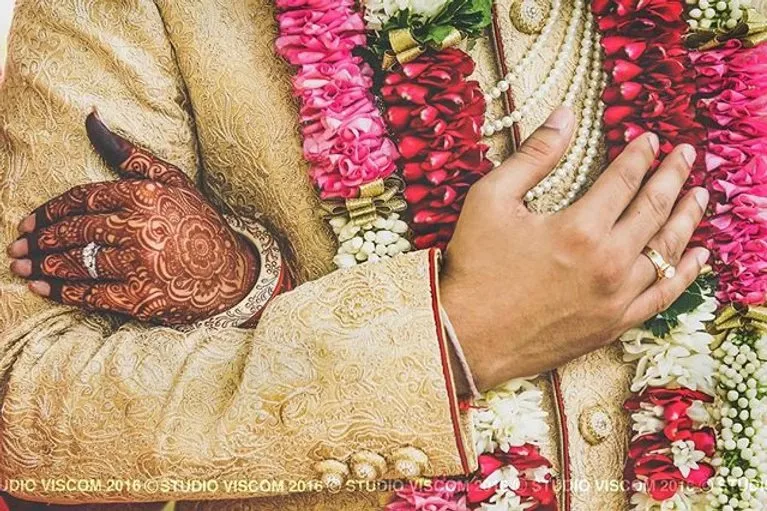 Bride and groom’s hands resting together over the wedding garlands, mehndi and rings visible